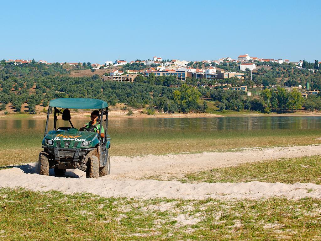 Lago Montargil&Villas Esterno foto The photo shows a small green all-terrain vehicle driving along a sandy path beside a body of water. In the background, there's a landscape with trees and rolling hills, along with a town or village visible on the hillside. The sky is clear, indicati