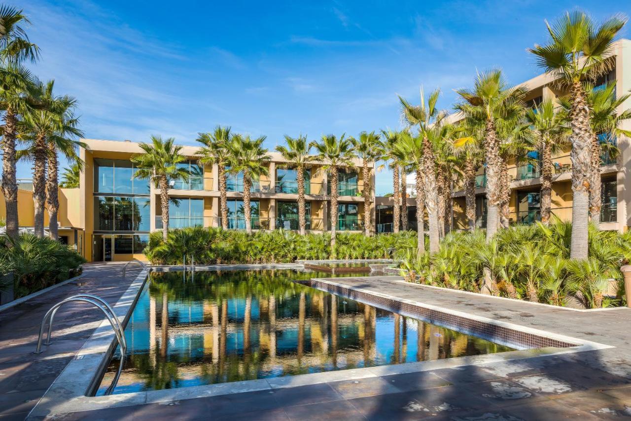 Lago Montargil&Villas Esterno foto The photo features a modern building surrounded by lush greenery and palm trees. In the foreground, there is a serene swimming pool with a reflective surface that captures the surrounding environment. The architecture of the building appears to have 