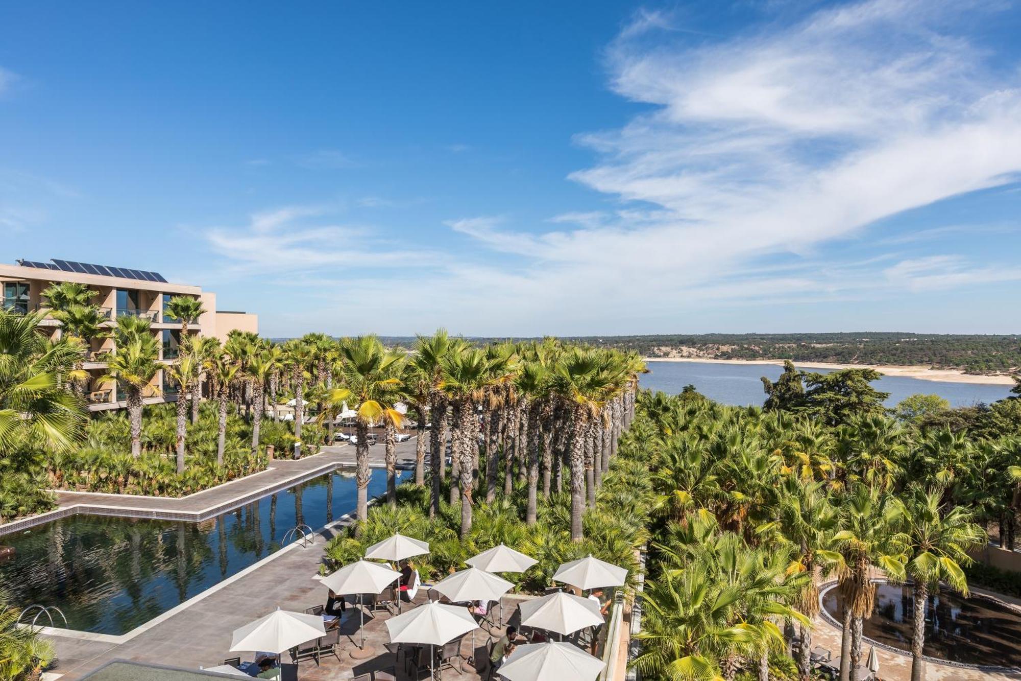 Lago Montargil&Villas Esterno foto The photo shows a scenic view of a resort or hotel area. In the foreground, there is a pool surrounded by palm trees and lounge chairs with umbrellas, suggesting a relaxing atmosphere. Beyond the pool, there is a body of water, likely a river or coas
