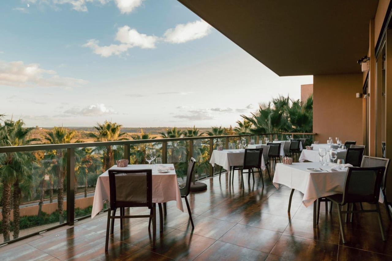 Lago Montargil&Villas Esterno foto The photo shows a dining area on a balcony with a view. There are several tables set for dining, each covered with white tablecloths. The chairs are wooden and have a simple design. In the background, there are palm trees and a scenic view of the lan