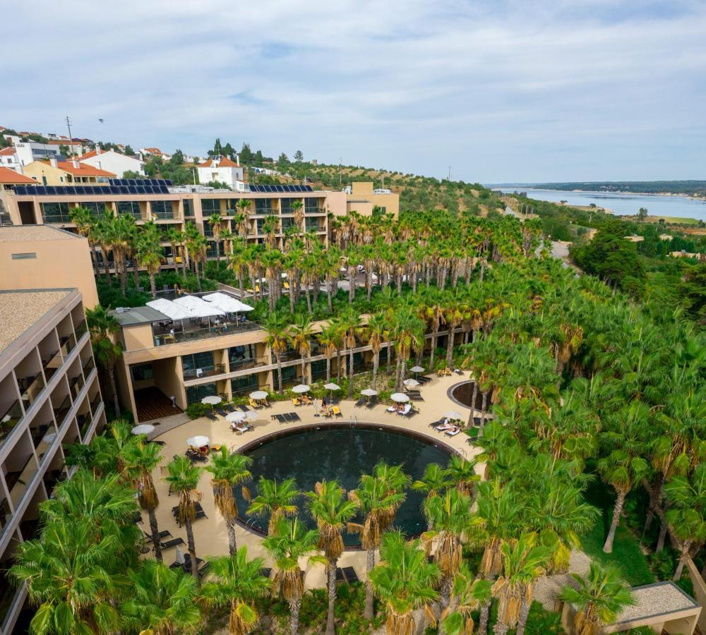 Lago Montargil&Villas Esterno foto The photo shows a modern hotel complex set amidst lush greenery, featuring numerous palm trees. In the foreground, there is a circular swimming pool surrounded by lounge chairs. The hotel itself has multiple levels with balconies overlooking the pool