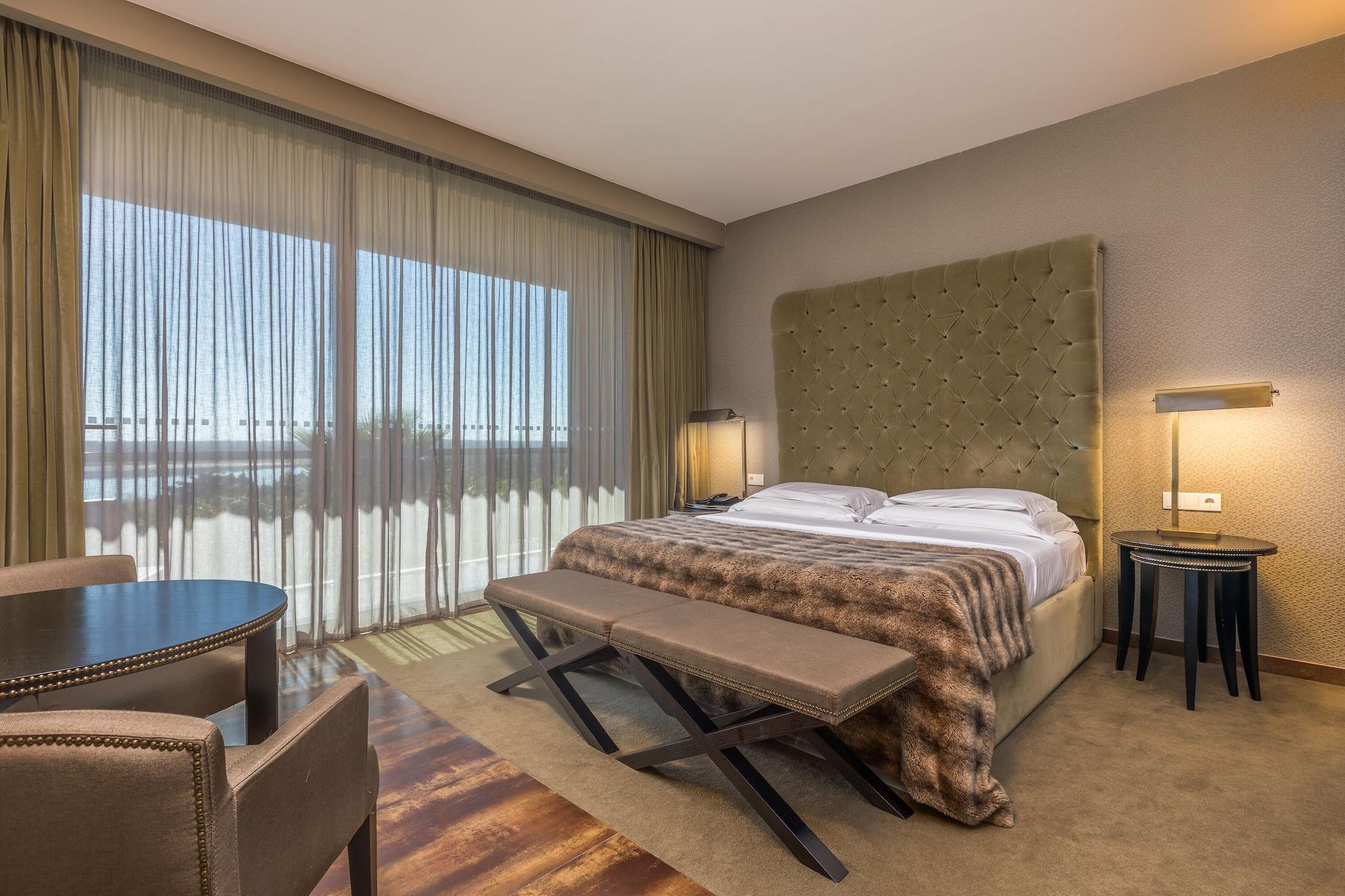 Lago Montargil&Villas Esterno foto The photo shows a modern hotel room featuring a large bed with a tufted headboard and a cozy blanket. There are curtains draped over large windows that allow natural light to flow in, providing a view of the outside. A small seating area includes two