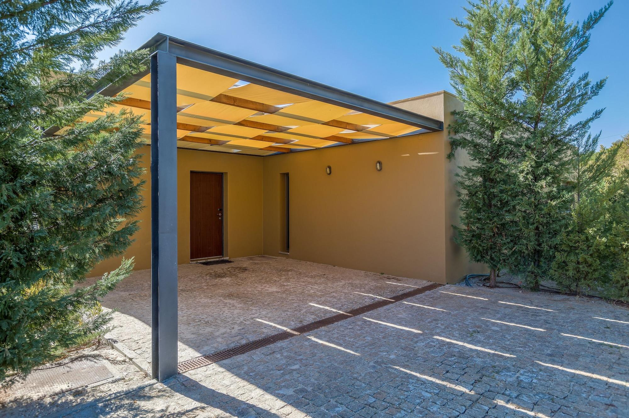 Lago Montargil&Villas Esterno foto The photo shows an exterior view of a modern structure with a carport. The carport has a sleek, metallic frame and a yellowish canopy above it. In front of the structure, there is a paved area made of stone or tiles. The main entrance features a wood