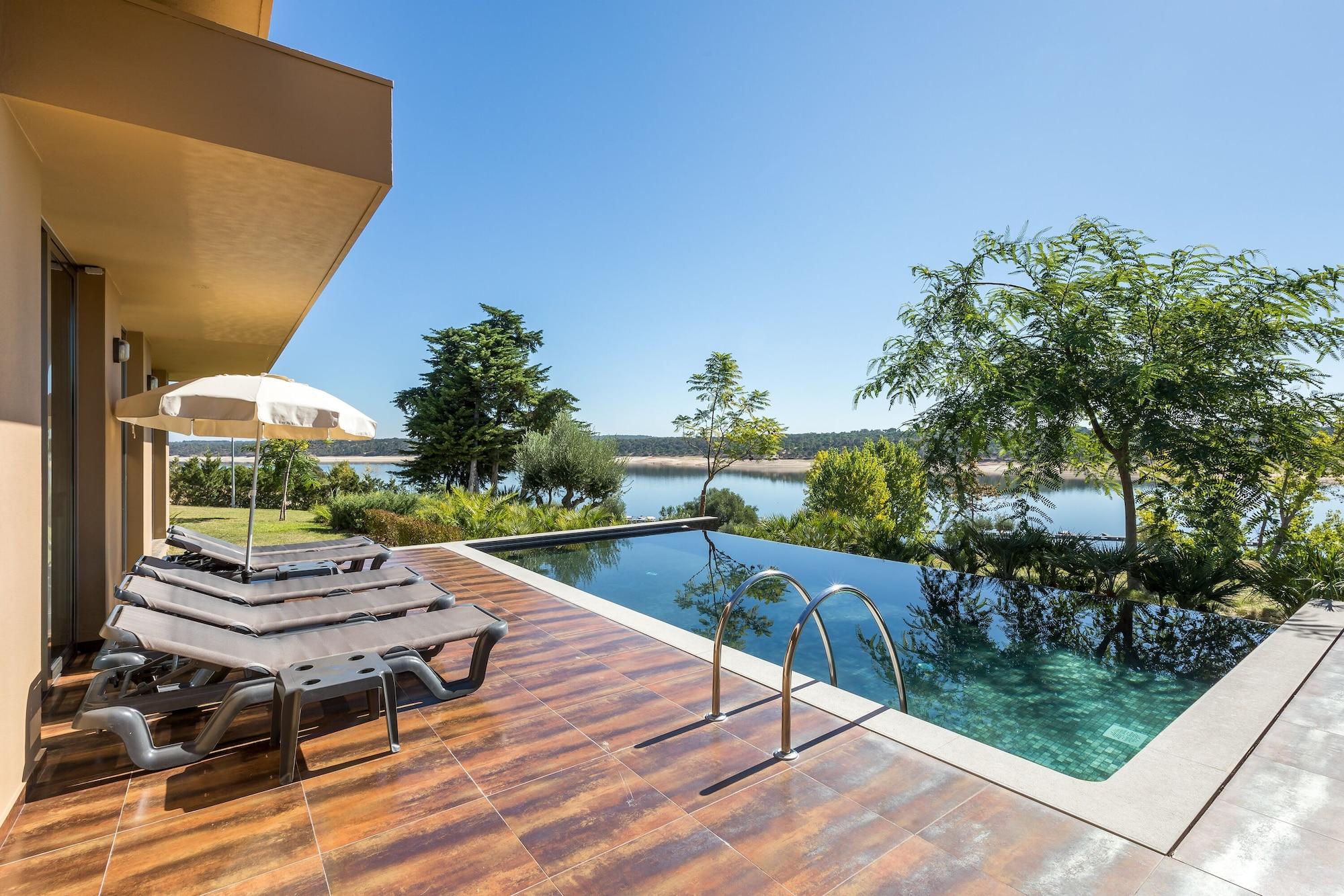 Lago Montargil&Villas Esterno foto The photo shows a luxurious outdoor setting by a pool. There are several lounge chairs arranged next to an infinity pool, which overlooks a scenic body of water and lush greenery. A parasol provides shade for relaxation. The sky is clear and blue, in