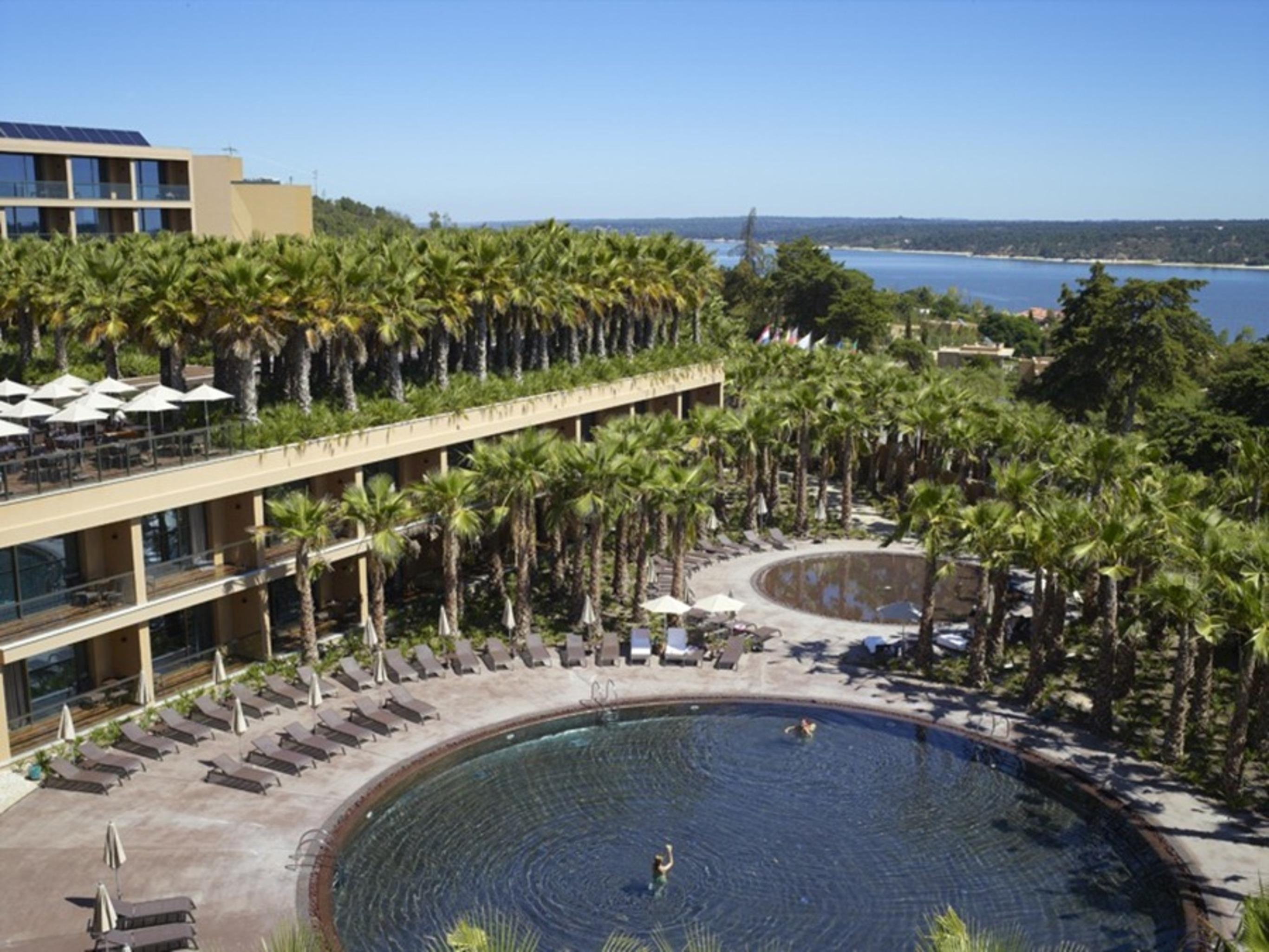 Lago Montargil&Villas Esterno foto The photo shows a scenic view of a resort area. In the foreground, there are circular pools surrounded by lounge chairs, with a few people enjoying the water. Palm trees line the edges, creating a tropical atmosphere. In the background, there is a mu