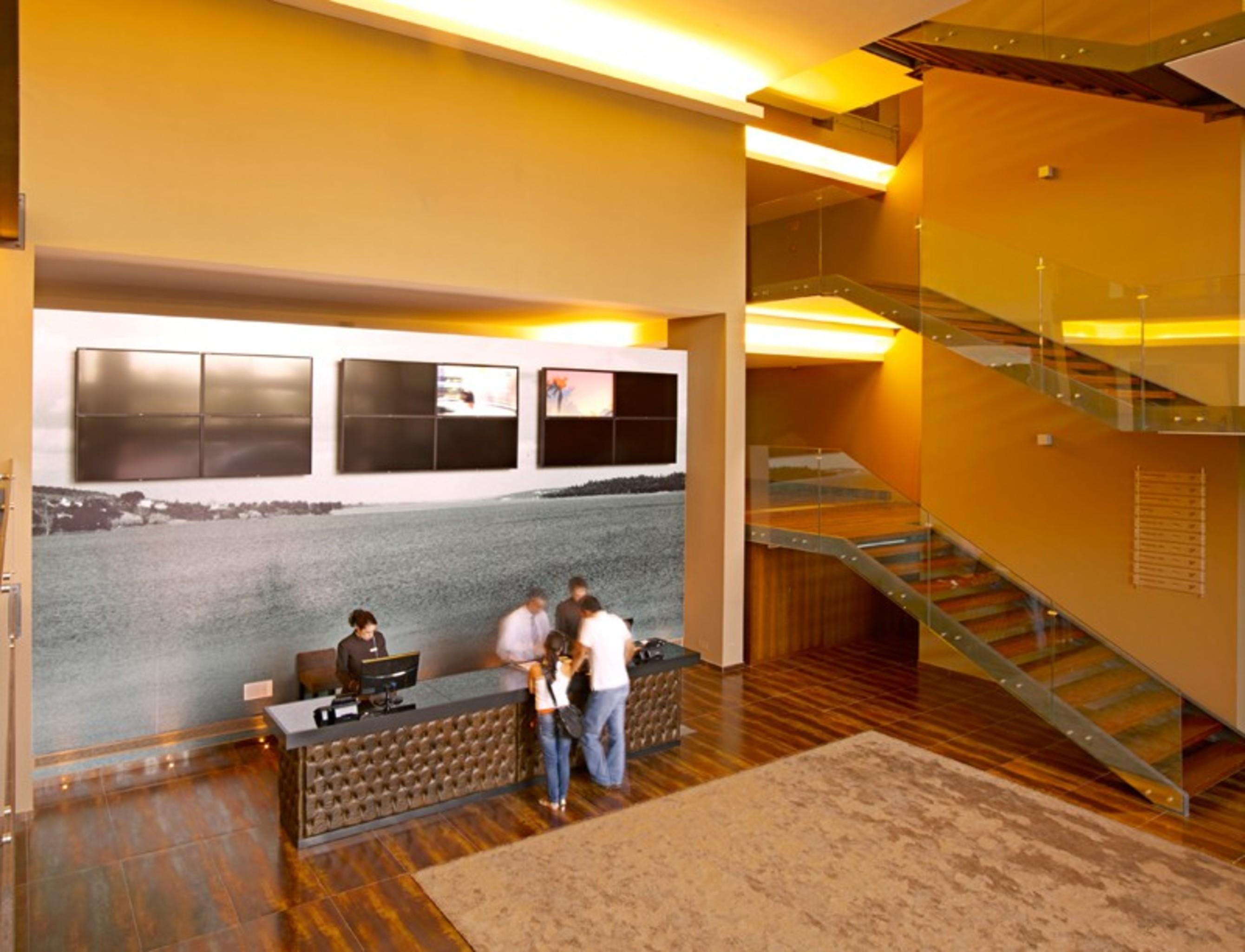 Lago Montargil&Villas Esterno foto The photo depicts a modern lobby area with a reception desk. The design features a combination of warm colors and wood flooring. In the background, there is a large wall-mounted screen or display, showcasing several monitors. The wall behind the desk