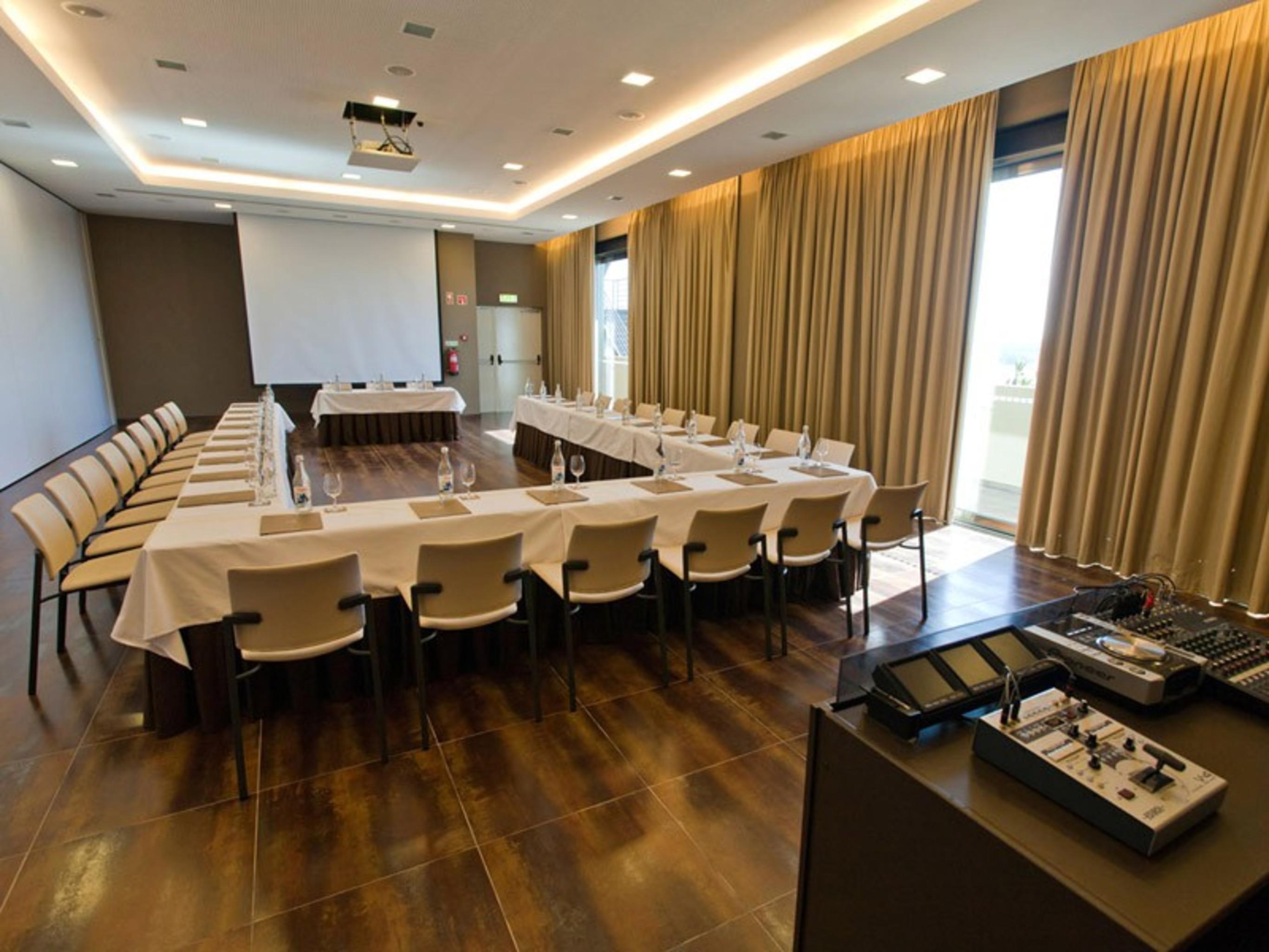 Lago Montargil&Villas Esterno foto The photo shows a modern conference room set up for a meeting. There are several tables arranged in a U-shape, surrounded by chairs. A large screen and projector are positioned at the front, with a table for speakers or presenters. The room has large