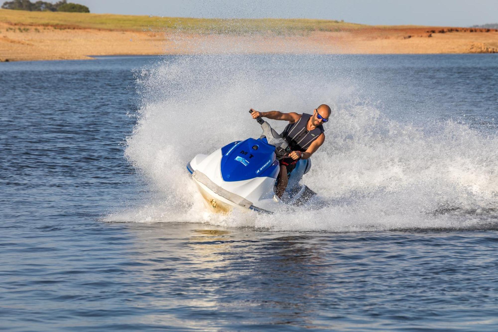 Lago Montargil&Villas Esterno foto The photo shows a person riding a jet ski over a body of water. The rider is wearing sunglasses and a life vest, leaning forward as the jet ski accelerates, creating a spray of water around them. The background features a natural landscape, likely a 