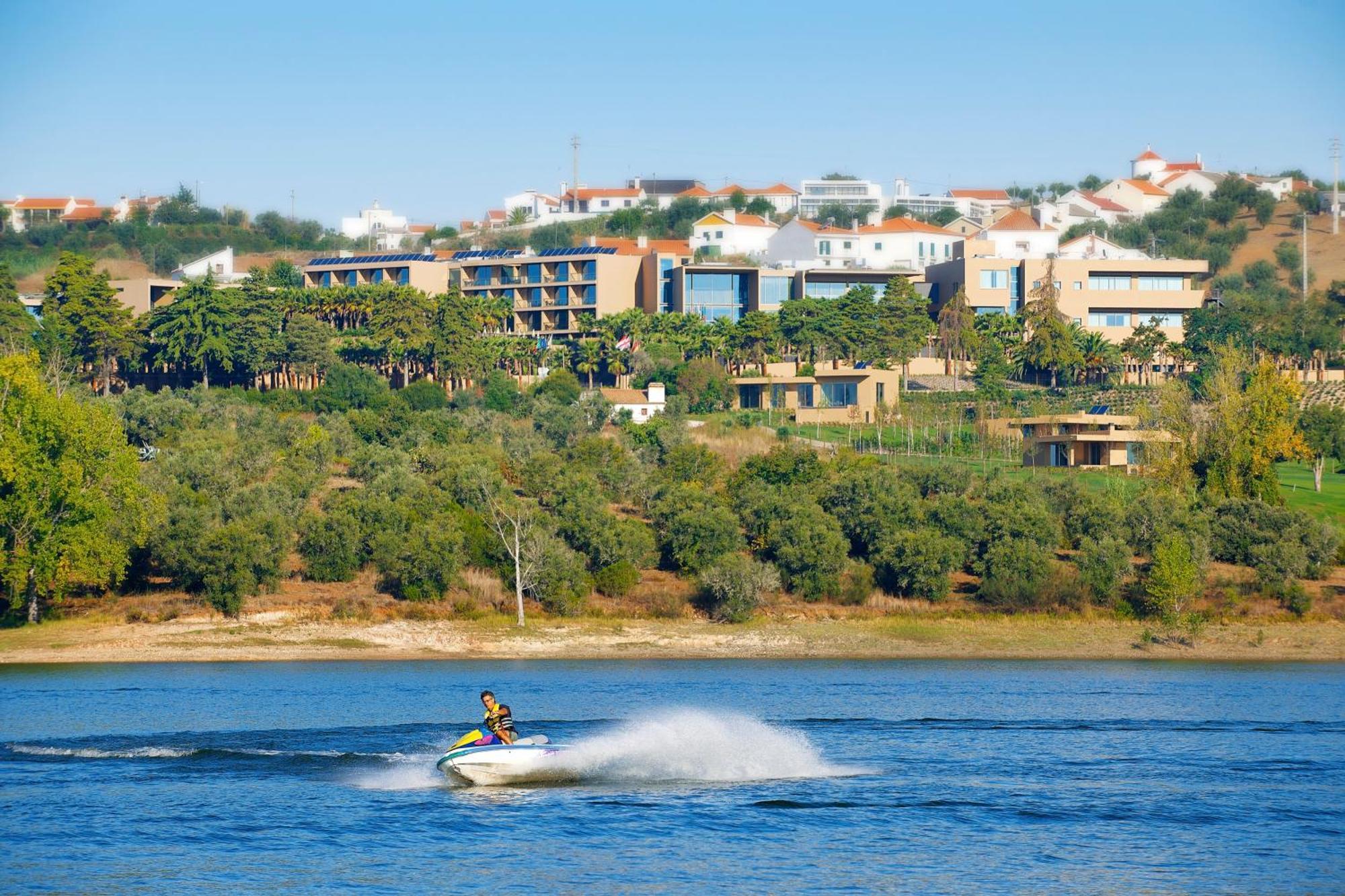 Lago Montargil&Villas Esterno foto The photo shows a person riding a jet ski on a body of water, creating a spray as they speed across the surface. In the background, there are modern buildings and a hillside with trees and greenery. The sky is clear and blue, suggesting a bright and 