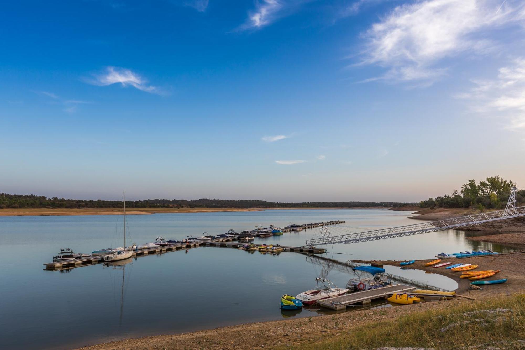 Lago Montargil&Villas Esterno foto The photo shows a serene lakeside scene. In the foreground, there are several boats moored at a dock, with some colorful kayaks and canoes lined up along the shore. The water is calm and reflects the sky, which features a few wispy clouds. In the bac