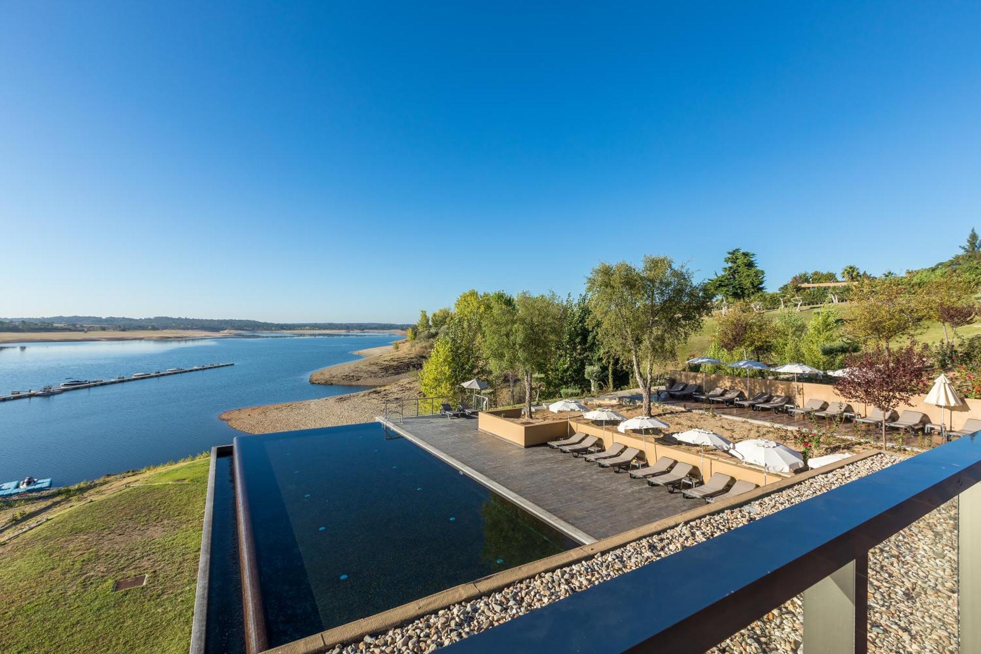 Lago Montargil&Villas Esterno foto The photograph depicts a scenic view of a tranquil water body, likely a lake or river, bordered by lush greenery. In the foreground, there is an infinity pool that blends seamlessly with the water beyond. Comfortable lounge chairs are arranged on a w