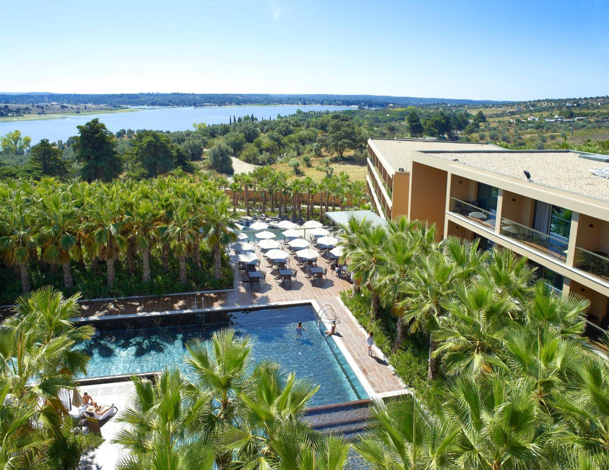 Lago Montargil&Villas Esterno foto The photo shows a scenic view of a modern hotel or resort. In the foreground, there is a swimming pool surrounded by palm trees, with lounge chairs positioned beside it. Sun umbrellas provide shade over the chairs. The background features a lush land