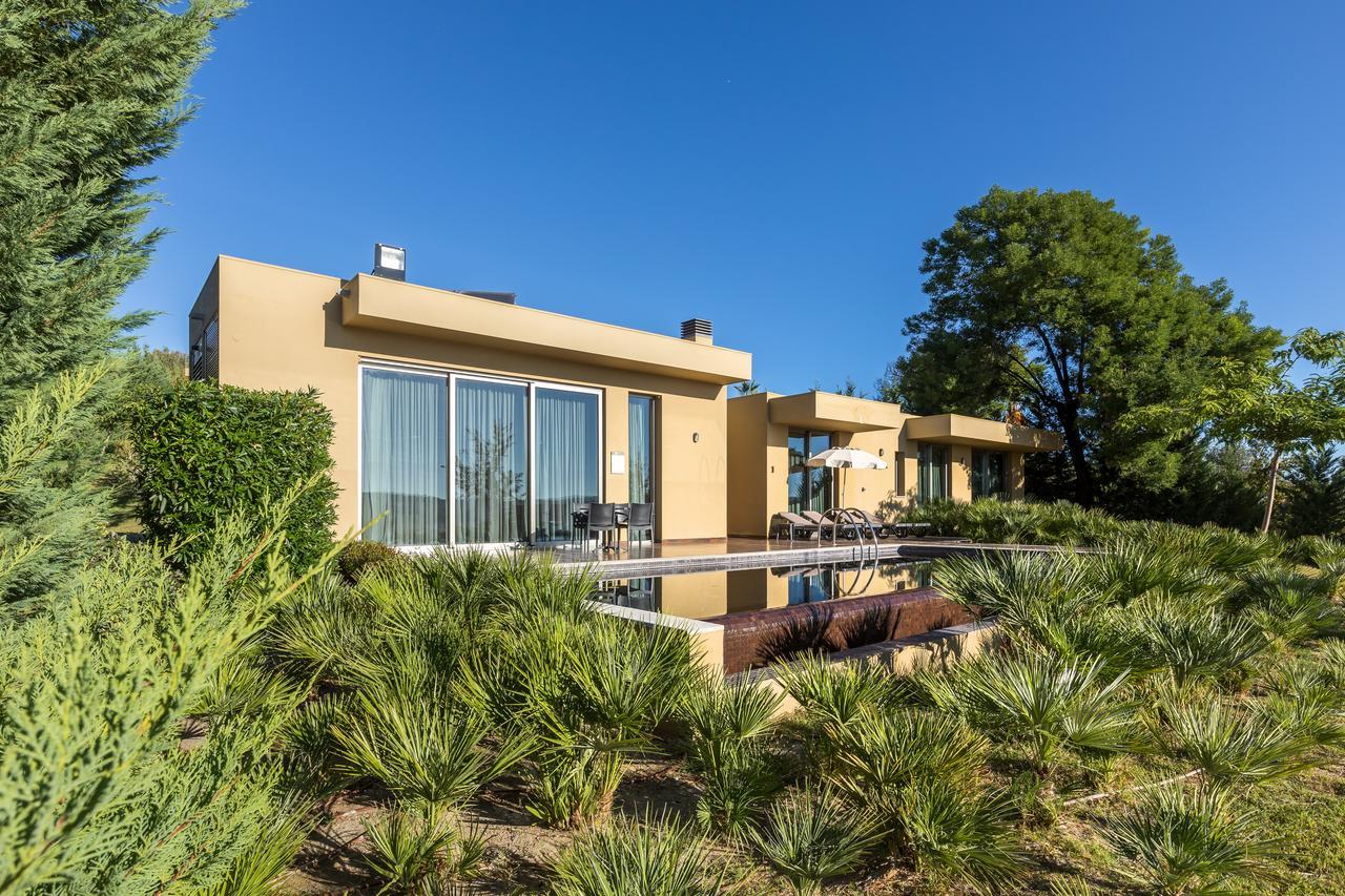 Lago Montargil&Villas Esterno foto The photo shows a modern villa or house set in a natural landscape. The building features a contemporary design with large glass windows and a beige exterior. In front of the house, there is a pool area, and the surrounding landscape includes greener