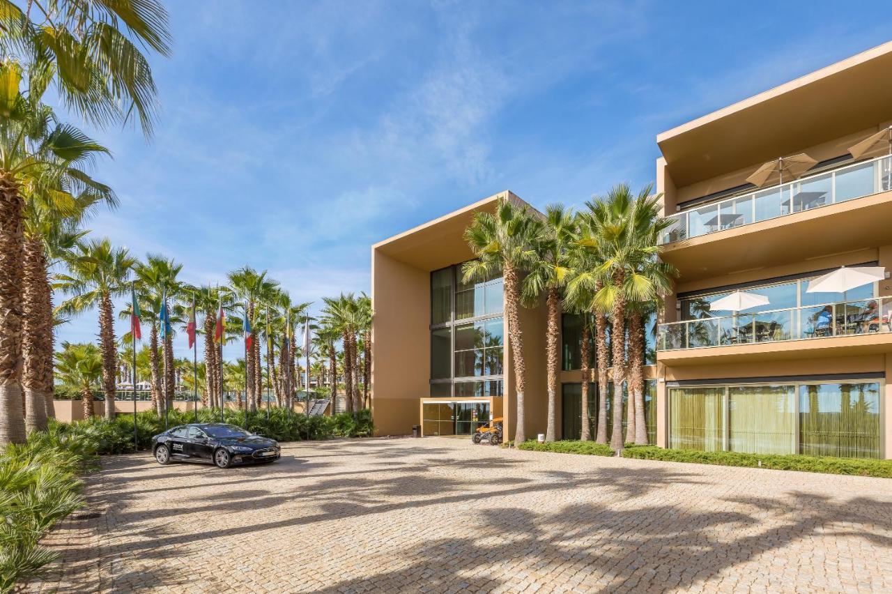 Lago Montargil&Villas Esterno foto The photo shows a modern building with a sleek design, featuring large glass windows and balconies. The structure is surrounded by a well-maintained outdoor area with palm trees, adding a tropical feel. In front of the building, there is a paved park