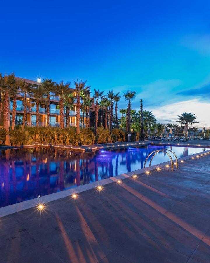 Lago Montargil&Villas Esterno foto The image shows a serene and inviting outdoor area with a large swimming pool that features a beautiful blue hue, likely illuminated by lights. Surrounding the pool are well-maintained palm trees and lush greenery, creating a tropical atmosphere. In 